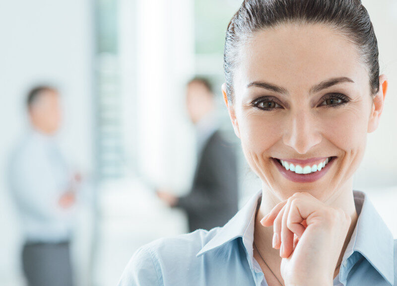 Confident beautiful smiling business woman in the office posing with hand on chin and looking at camera office interior and business team on background selective focus